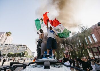 Two protesters wave Mexican flags while standing on a vandalized Waymo vehicle during a demonstration in Los Angeles, California, on June 8, 2025, following a series of aggressive federal immigration operations in the city.
